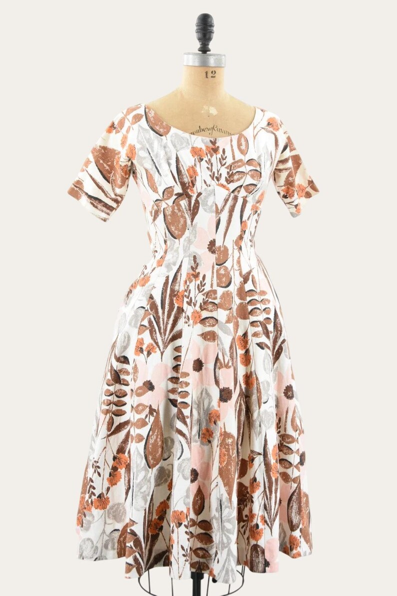 May include: A vintage white dress with a floral pattern in shades of brown, orange, and pink. The dress features short sleeves and a fitted bodice that flares into a full skirt. The dress is displayed on a mannequin.
