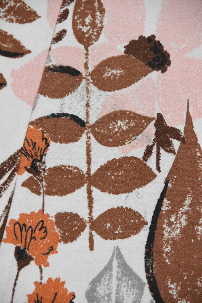 May include: Close-up of a fabric with a floral pattern. The design features brown, orange, and black floral and leaf motifs against a white background. Pink, gray, and brown shapes are also present, creating a textured look.