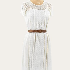 May include: A vintage white dress with a crochet yoke and short sleeves. The dress features a textured pattern and is cinched at the waist with a brown leather belt. The dress is displayed on a mannequin.
