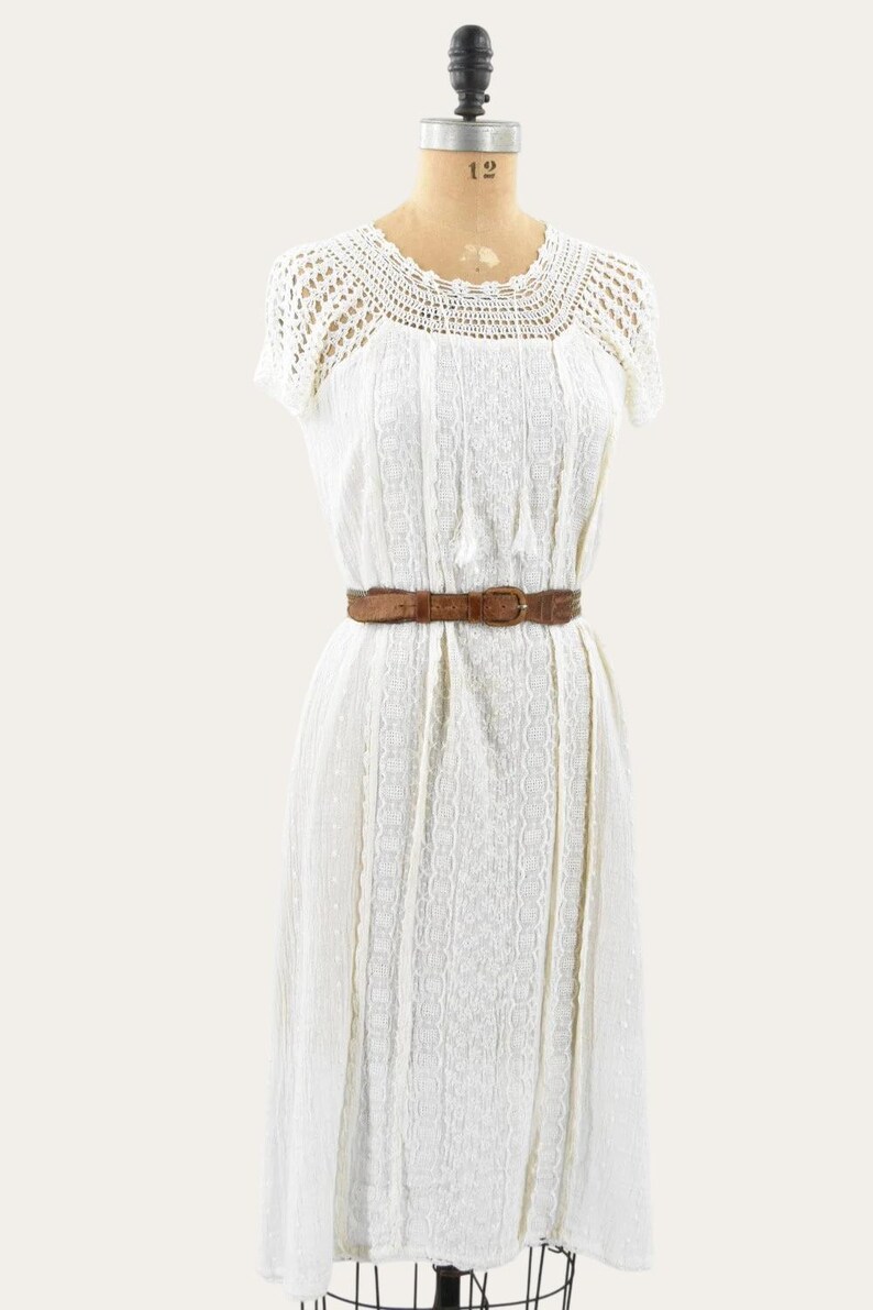 May include: A vintage white dress with a crochet yoke and short sleeves. The dress features a textured pattern and is cinched at the waist with a brown leather belt. The dress is displayed on a mannequin.