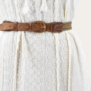 May include: A close-up of a white dress with a brown leather belt. The belt has a rectangular buckle and a woven section. The dress has a textured pattern and decorative tassels.