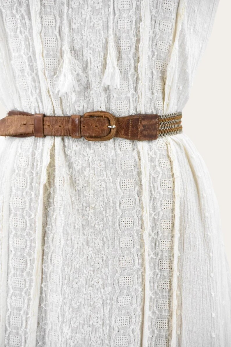May include: A close-up of a white dress with a brown leather belt. The belt has a rectangular buckle and a woven section. The dress has a textured pattern and decorative tassels.