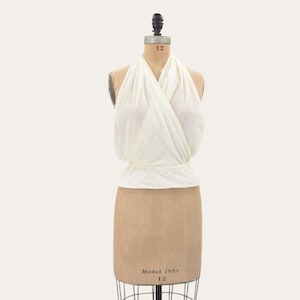 May include: A white sleeveless halter top with a draped, wrap-style bodice and a gathered waistline. The garment is displayed on a beige dress form mannequin that features the text Model 1953 and the number 12 printed on the torso.