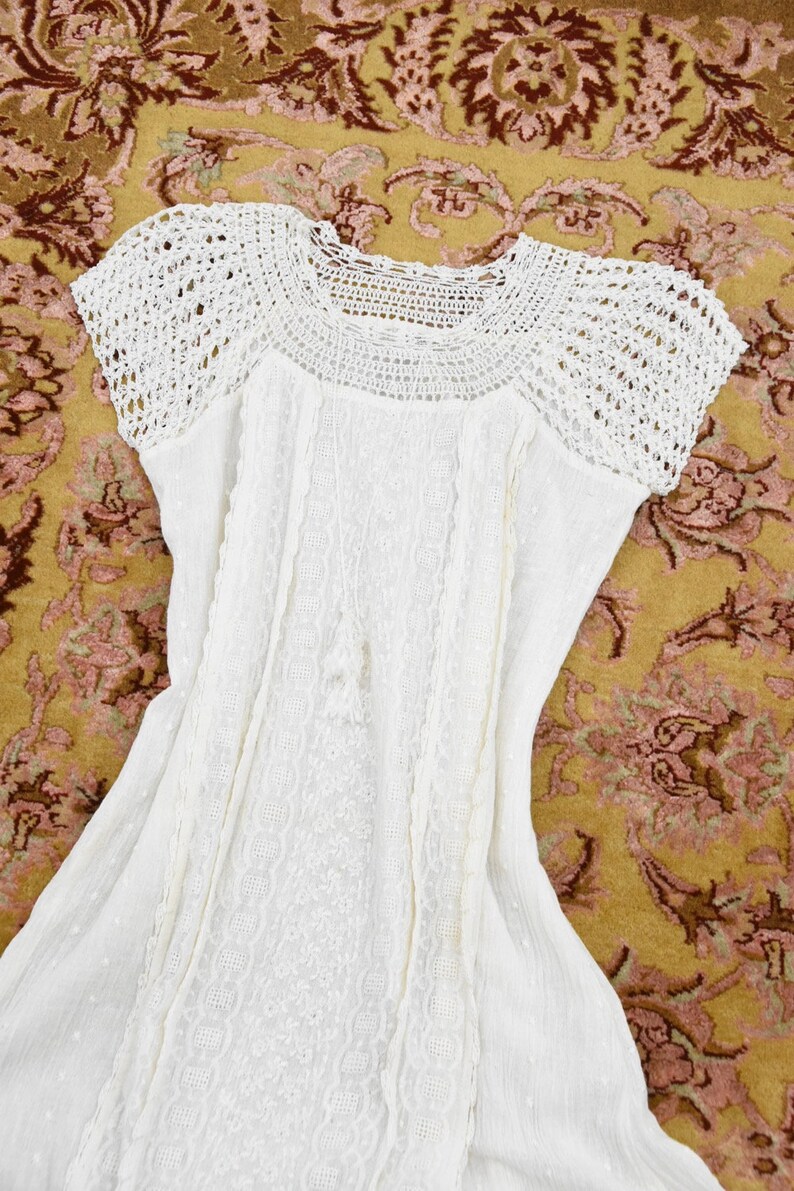 May include: White dress with a crochet yoke and short sleeves. The dress features detailed embroidery and a flowing skirt. The garment is made of a lightweight fabric, perfect for warm weather. The dress is laid out on a patterned rug.