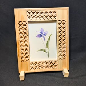 Puede incluir: Un marco rectangular de madera clara con intrincados patrones de celosía cortados con láser en los cuatro lados. El marco sostiene una pieza blanca que muestra una acuarela de una sola flor de violeta morada con hojas verdes.