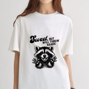 Könnte beinhalten: Ein weißes T-Shirt mit kurzen Ärmeln und einer schwarzen grafischen Illustration eines Waschbären mit erhobenen Pfoten. Über dem Waschbären steht der Text "Sweet, BUT WILL THROW HANDS" in einer fetten, stilisierten schwarzen Schrift.