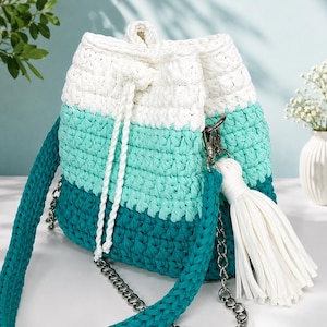 Könnte beinhalten: Eine Beuteltasche aus gehäkeltem Garn mit einem horizontalen Farbblock-Design in Weiß, Mintgrün und Dunkelblaugrün. Die Tasche verfügt über einen weißen Kordelzug, eine passende weiße Quaste und einen Riemen aus gehäkeltem Garn und silberner Kette.