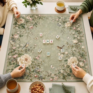 May include: A square mahjong mat featuring a sage green background with detailed illustrations of white and pink peonies, small birds, and butterflies. Several white mahjong tiles with red, green, and black characters are scattered across the surface.