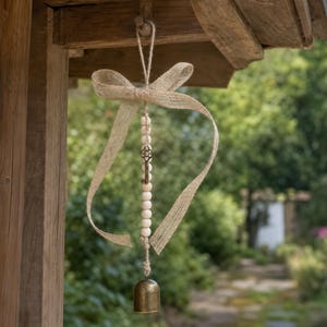 May include: A hanging ornament featuring a rustic burlap ribbon tied in a bow at the top, a vertical strand of light-colored wooden beads, a small metal skeleton key, and a brass-toned bell suspended from the bottom of the bead strand.