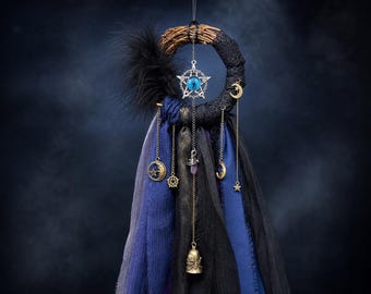 May include: A circular wall hanging featuring a woven twig base wrapped in black lace and fabric. It includes a central silver pentagram charm with a blue eye, black feathers, and hanging chains with gold-toned moon, star, and skull bell charms.