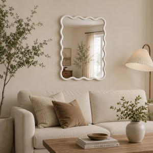 May include: A rectangular wall mirror with a white, wavy, scalloped frame. The mirror is mounted on a beige wall above a light-colored sofa. The reflective surface shows a room interior with a window, curtains, and a framed picture on the wall.