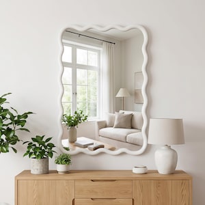 May include: A rectangular wall mirror featuring a thick, white, wavy frame. The mirror has rounded corners and a continuous undulating border design. The reflective surface shows a room interior with a window, a sofa, and a lamp.