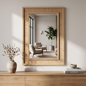 May include: A rectangular wall mirror featuring a light-toned wooden frame with an inset panel of woven cane webbing. The mirror reflects a room containing two wooden armchairs, a potted plant, and a wall sconce, all set against a plain wall.