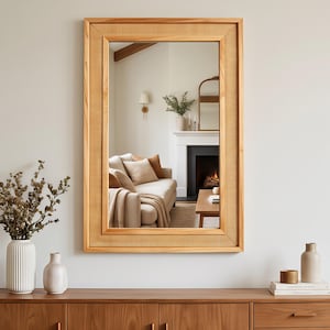 May include: A rectangular wall mirror featuring a light-toned wooden frame with a woven cane border. The mirror reflects a living room interior with a sofa, a fireplace, and decor. The frame has a smooth finish and a clean, structured design.