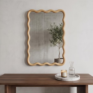 May include: A rectangular wall mirror featuring a light-toned wooden frame with a continuous wavy, scalloped edge. The mirror reflects a potted plant, while a marble tray holding a glass carafe and a lit candle sits on the wooden surface below.