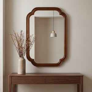 May include: A rectangular wall mirror with a dark brown wooden frame featuring rounded corners and decorative indentations. The mirror reflects a hanging pendant light with a light gray shade and a wooden top, plus a textured beige vase.