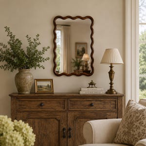 May include: A rectangular wall mirror with a dark brown wavy frame hanging above a wooden sideboard. The sideboard features two drawers and two cabinet doors with black knobs. A lamp with a pleated shade and a vase with greenery sit on top.