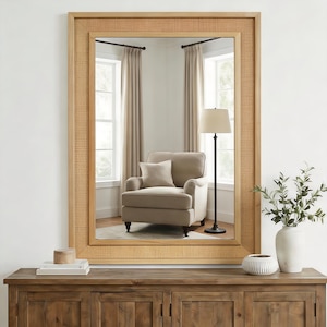 May include: A rectangular wall mirror featuring a light-toned wooden frame with an inset border of woven cane webbing. The mirror reflects a room interior containing a beige armchair, a floor lamp, and curtains, providing a clear view.