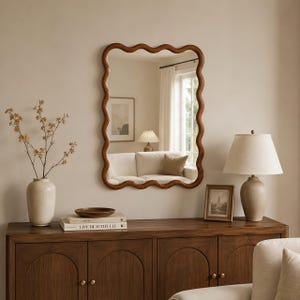 May include: A rectangular wall mirror with a wavy, scalloped wooden frame. The mirror is mounted above a wooden sideboard that holds a ceramic vase with branches, a small bowl, two stacked books, a framed picture, and a table lamp.