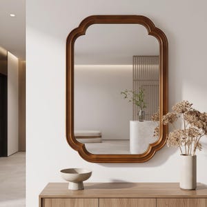 May include: A rectangular wall mirror with rounded, scalloped corners and a smooth, medium-toned wooden frame. The mirror reflects a bright interior space with a white marble pedestal, a green plant, and a light-colored bench in the background.
