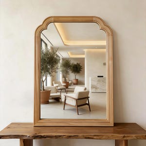 May include: A rectangular wall mirror with a light-toned wooden frame featuring a curved, arched top design. The mirror reflects a bright interior space with cream-colored armchairs, potted olive trees, and a reception desk area.