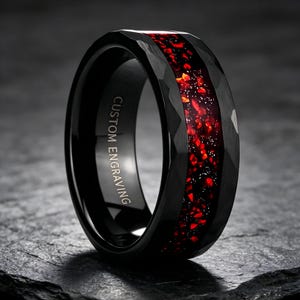 Puede incluir: Un anillo negro con una superficie exterior facetada y una incrustación central de fragmentos de piedra roja y motas plateadas brillantes. La banda interior es lisa y muestra claramente el texto grabado CUSTOM ENGRAVING.