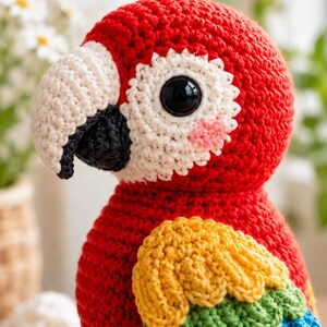 May include: A handcrafted crochet parrot toy featuring a vibrant red head and body, a large cream-colored beak with a black tip, and a white eye patch surrounding a black plastic eye. The wing displays layered yellow, green, and blue yarn feathers.