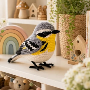 May include: A handcrafted crochet bird figurine featuring a grey head and back, a bright yellow throat and chest, and a white belly. The bird has black wing markings with white accents, a small black beak, and thin black legs standing on a surface.