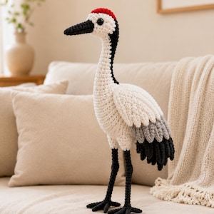 May include: A crocheted crane figurine featuring a white body, a black neck and beak, and a bright red patch on the top of its head. The bird has gray and black wing feathers and long, thin black legs, all crafted with a visible yarn texture.
