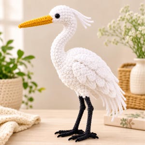 Peut inclure: Une figurine de héron crochetée à la main avec un corps blanc au plumage texturé, un long bec jaune et des pattes noires. L'oiseau possède un petit œil noir et une crête de plumes en fil blanc sur la tête, se tenant debout sur une surface plane.
