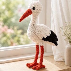 Puede incluir: Una figura de cigüeña tejida a ganchillo a mano con cuerpo blanco, puntas de alas negras y un pico largo y puntiagudo de color naranja. El ave se sostiene sobre dos patas naranjas delgadas con pies palmeados y tiene ojos negros redondos.