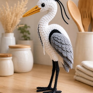 Peut inclure: Une figurine de héron crochetée à la main avec un corps blanc, des ailes grises et de longues pattes noires. L'oiseau possède un bec jaune vif, une calotte noire et deux fins brins décoratifs noirs partant de l'arrière de la tête en laine.