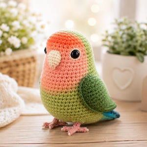 Op de afbeelding: Een gehaakt dwergpapegaai-figuurtje met een verloop van perzik en oranje op de kop, een lichtgroene borst en donkergroene vleugels. De vogel heeft zwarte plastic ogen, een crèmekleurige snavel en kleine roze pootjes, gemaakt met zichtbare steken.