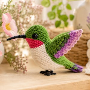 Puede incluir: Una figura de colibrí tejida a ganchillo con cabeza y espalda verdes, pecho blanco, garganta de color rosa brillante y alas con puntas moradas. El ave tiene un pico negro largo y fino y ojos negros pequeños, apoyada sobre diminutas patas negras.
