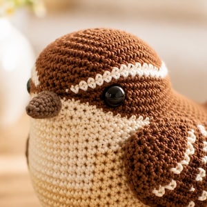 Könnte beinhalten: Eine handgefertigte Häkelvogel-Figur mit einem abgerundeten Körper aus braunem und cremefarbenem Garn. Der Vogel hat einen kleinen braunen Schnabel, zwei glänzende schwarze Perlenaugen und aufwendige weiße Stickdetails an Kopf und Flügeln.