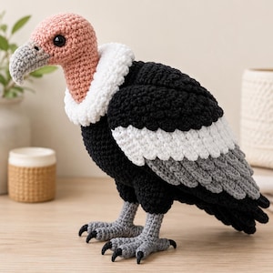 May include: A crocheted vulture figurine featuring a light pink head, a grey beak, and a white ruff around the neck. The body is primarily black with white and grey wing feathers, and the bird stands on two grey crocheted legs with black claws.