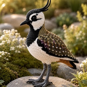 May include: A crocheted bird figurine featuring a black head with a prominent crest, a white face patch, and a dark beak. The body displays a mix of olive green, brown, and white yarn textures, with long grey crocheted legs and feet standing upright.