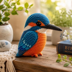 Pode incluir: Uma figura de pássaro guarda-rios em croché com cabeça e asas azul-brilhante, peito cor de laranja, uma mancha branca no pescoço e um bico comprido preto. O pássaro tem pequenos olhos de contas pretas e um ponto detalhado no corpo.