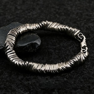 Könnte beinhalten: Ein silberfarbenes Armband, das aus zahlreichen dünnen, flachen, rechteckigen Metallsegmenten besteht, die zu einer durchgehenden kreisförmigen Kette verbunden sind. Das Armband hat eine metallische Oberfläche und einen Karabinerverschluss.