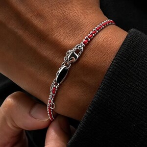 May include: A bracelet featuring a vibrant red cord intertwined with a polished silver-toned chain. The design includes a central metallic clasp element with a smooth black inlay, creating a contrast between the bright red and the metallic silver.