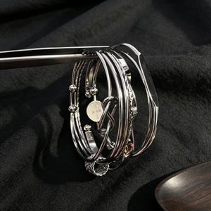 May include: A set of silver-toned metal bracelets featuring various designs, including a smooth band, a twisted rope texture, a small infinity knot detail, and a circular charm with engraved text, all held together by a pair of metal tweezers.
