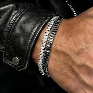 May include: Two tennis bracelets worn together on a wrist. One bracelet features clear, baguette-cut stones set in a silver-toned metal, while the other bracelet features black, baguette-cut stones set in a matching silver-toned metal frame.