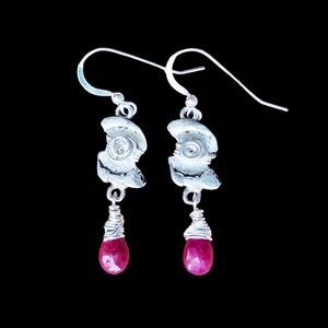 May include: A pair of silver earrings. Each earring has a textured, abstract silver shape with a coiled wire detail and a dangling, faceted, teardrop-shaped, deep red gemstone. The earrings hang from silver ear wires. The background is black.
