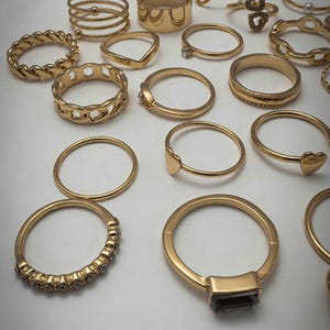May include: A collection of gold-toned rings in various designs, including plain bands, twisted textures, heart shapes, chain links, and one ring featuring a dark rectangular stone and another with a row of small clear stones.