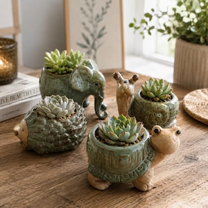 May include: A collection of four ceramic animal-shaped planters in shades of sage green and tan. The set includes an elephant, a snail, a hedgehog, and a turtle, each featuring a small succulent plant nestled within its hollow back or shell.