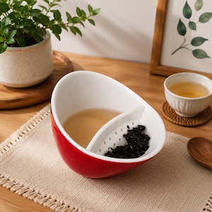 Puede incluir: Un cuenco de té de cerámica roja con interior blanco y un divisor perforado integrado. El cuenco contiene té líquido claro en un lado y hojas de té oscuras en el otro, separados por la pared de filtro de cerámica blanca integrada.