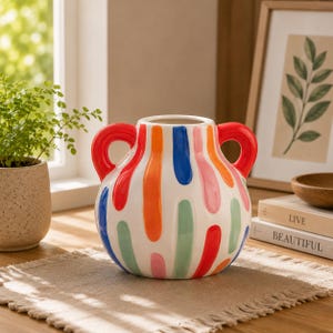 Può includere: Un vaso in ceramica rotondo con due manici rossi, caratterizzato da una base bianca decorata con pennellate verticali nei toni dell'arancione, blu, rosa e verde. Il vaso ha un corpo largo e un'apertura stretta nella parte superiore.