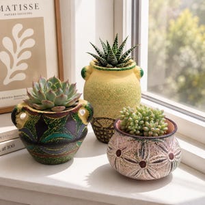 Puede incluir: Tres macetas de cerámica con plantas suculentas. Una es de color verde oscuro y negro con un patrón geométrico, otra es amarilla con acabado texturizado y la tercera es rosa claro con un diseño floral oscuro. Cada maceta tiene dos asas.