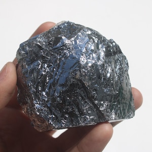 Puede incluir: Un trozo bruto y dentado de metal de silicio con una superficie metálica de color gris oscuro altamente reflectante. El espécimen mineral presenta bordes afilados e irregulares y una textura cristalina que capta la luz, creando reflejos brillantes.