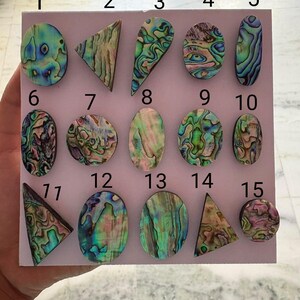Op de afbeelding: Een verzameling van vijftien gepolijste abalone schelp cabochons gerangschikt op een lichtroze bord, genummerd van 1 tot 15. De stukken vertonen iriserende patronen in tinten groen, blauw, paars en roze, met diverse vormen zoals ovalen, driehoeken en cirkels.